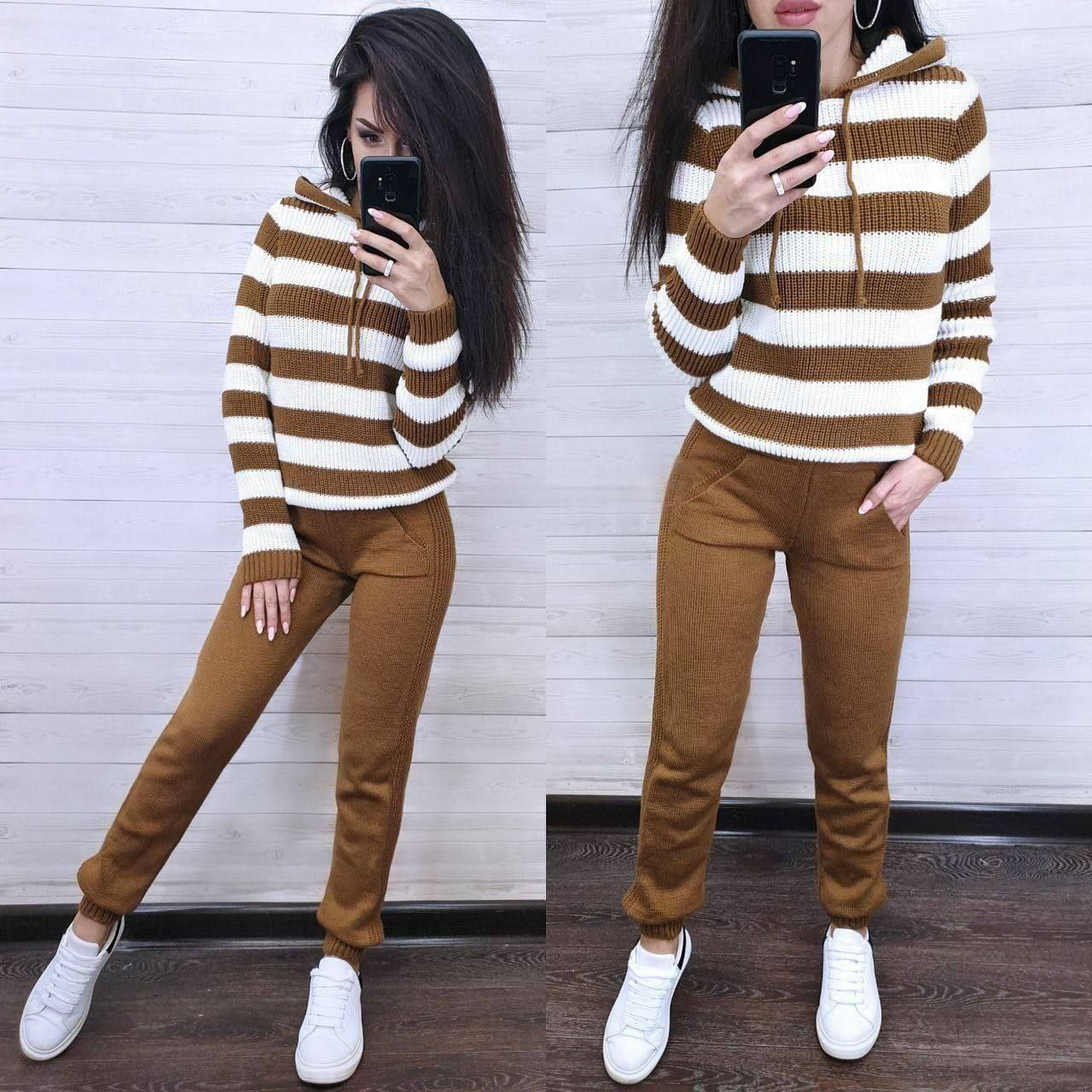 Woman wearing a brown and white striped sweater and brown pants, standing against a white wall.