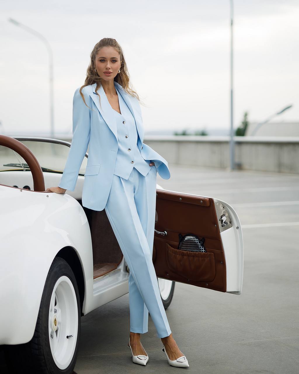 Woman in a light blue suit standing next to a vintage car with an open door.