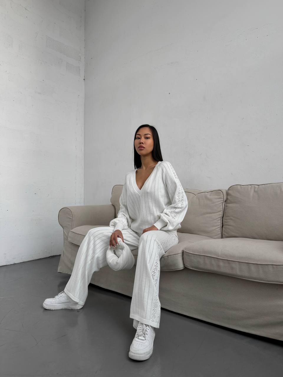Woman in a white outfit sitting on a beige sofa against a plain wall.