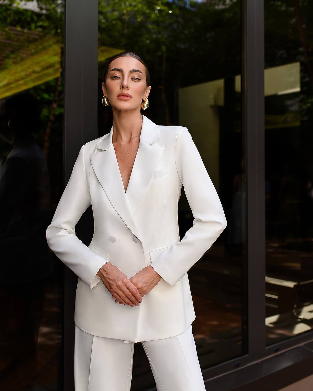 Woman wearing a white suit standing outdoors with greenery in the background