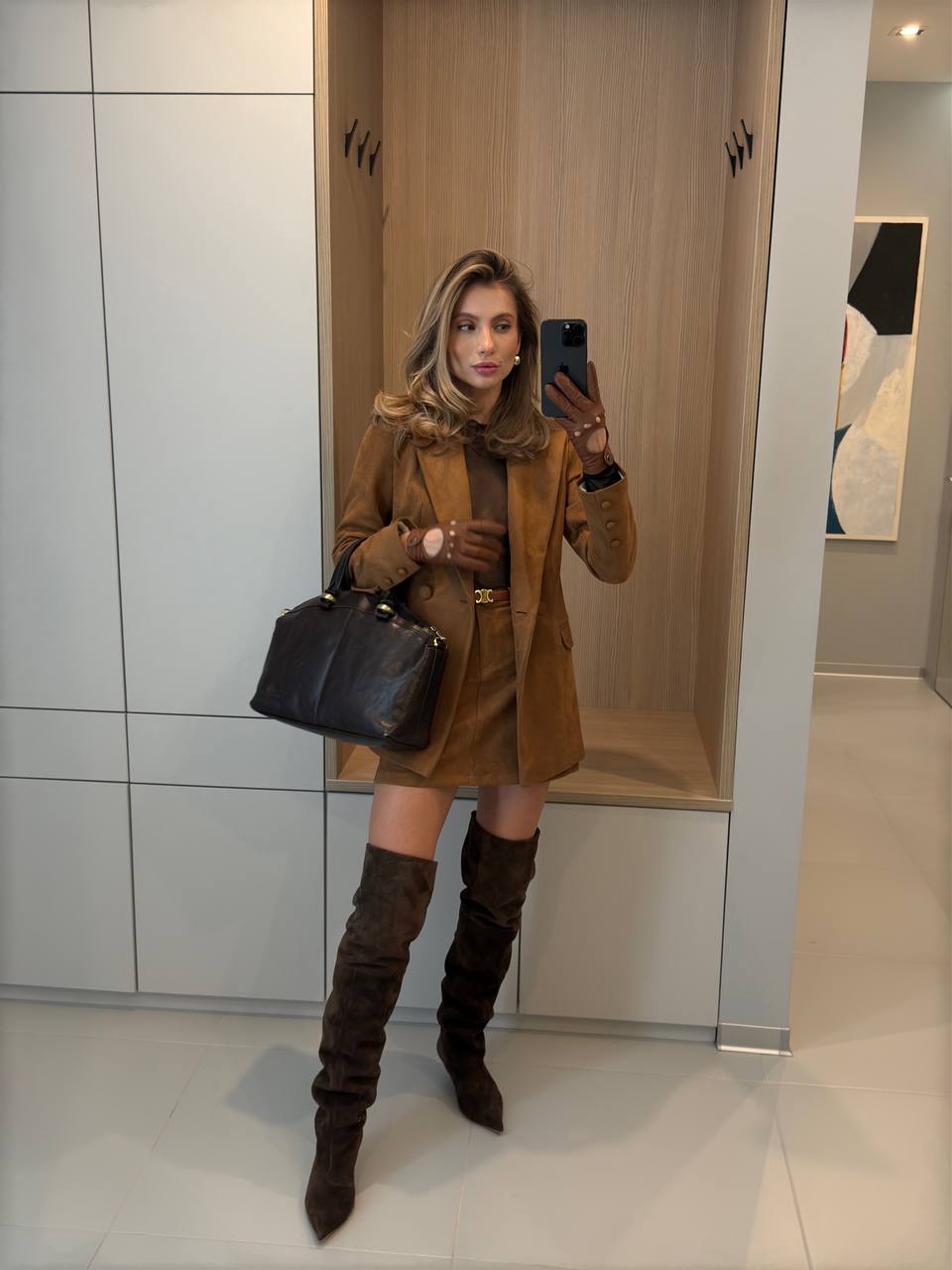 Woman taking a mirror selfie wearing a brown coat and knee-high boots in a modern interior setting.
