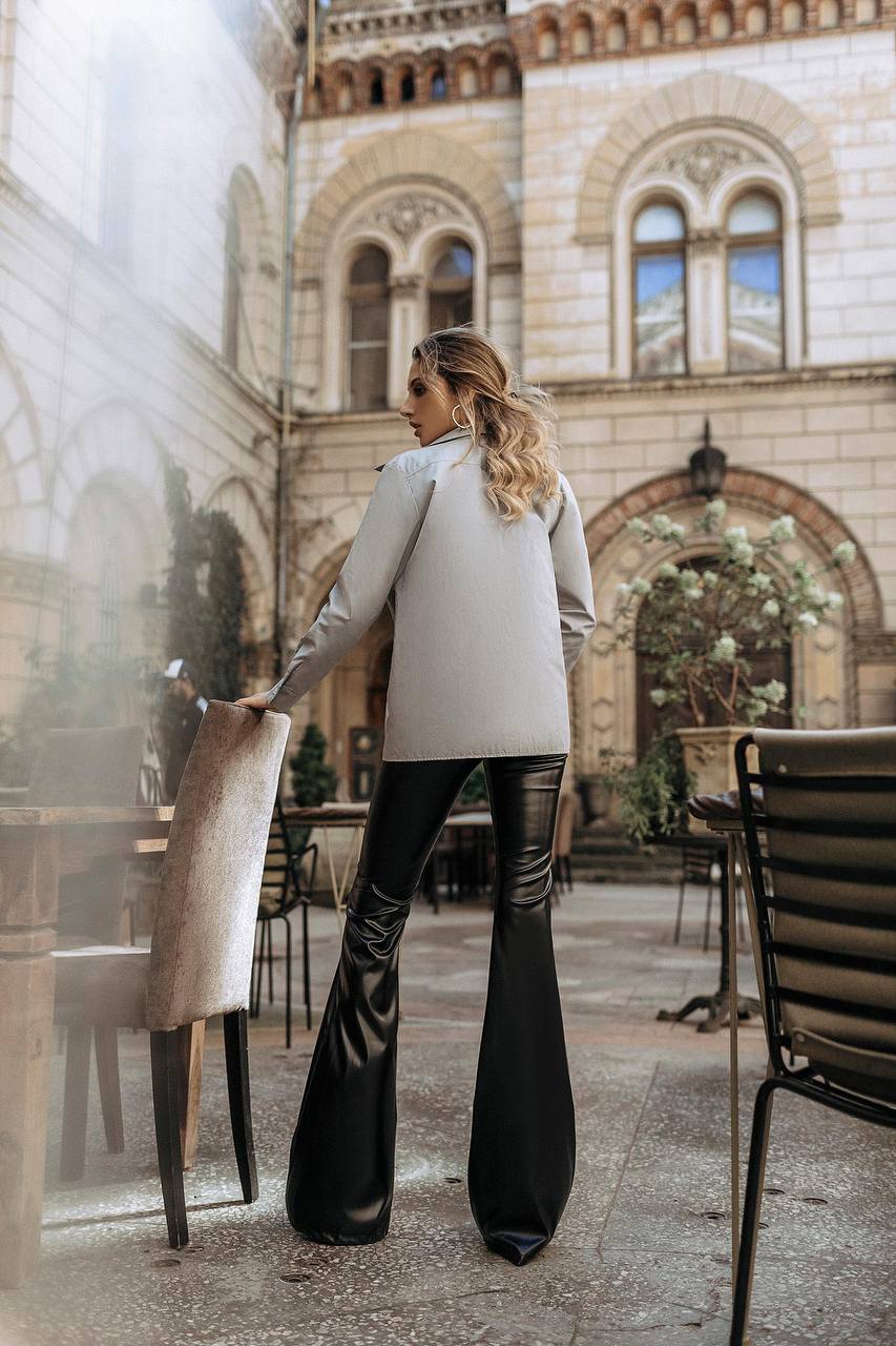 Woman in a beige coat and black flared pants standing in an outdoor setting with architectural elements.