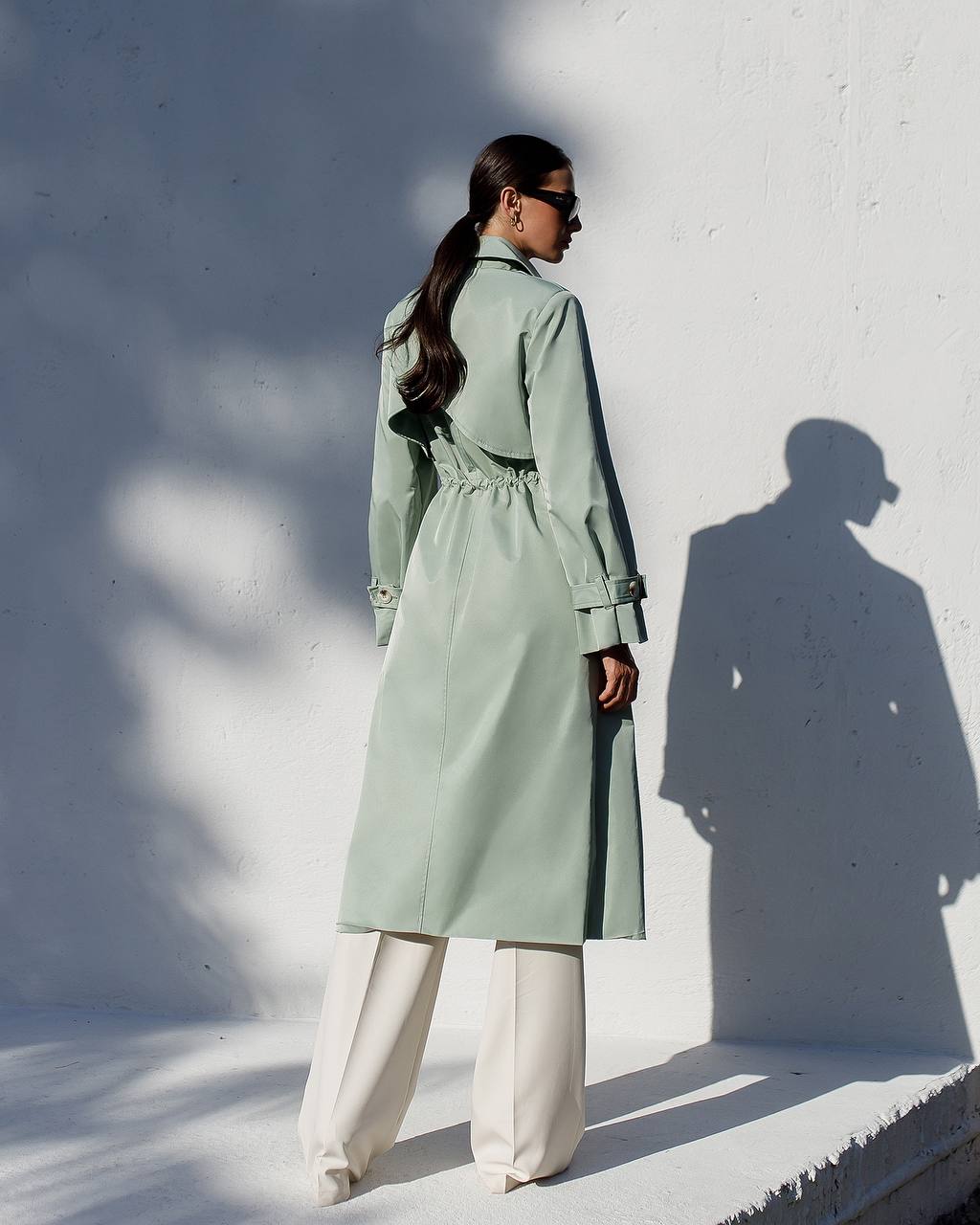 Person wearing a light green trench coat against a white wall with shadows.