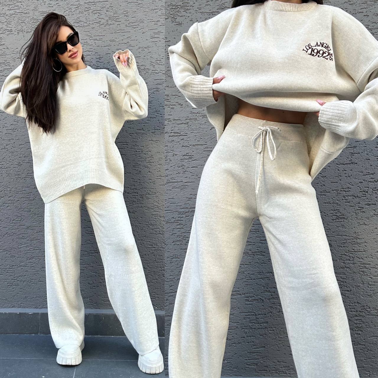 Woman wearing a beige sweater and pants set against a gray wall.