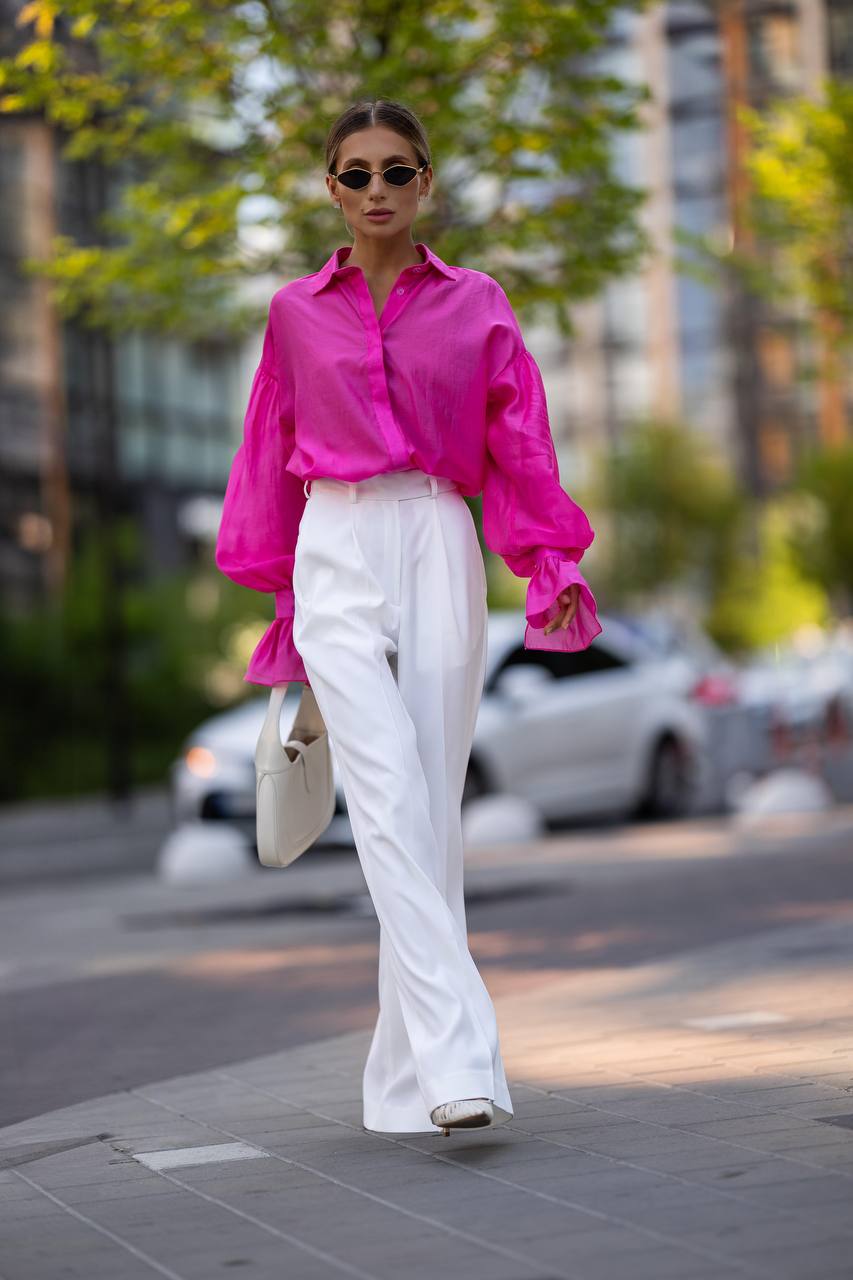 Woman wearing a bright pink shirt and white pants walking on a city street.