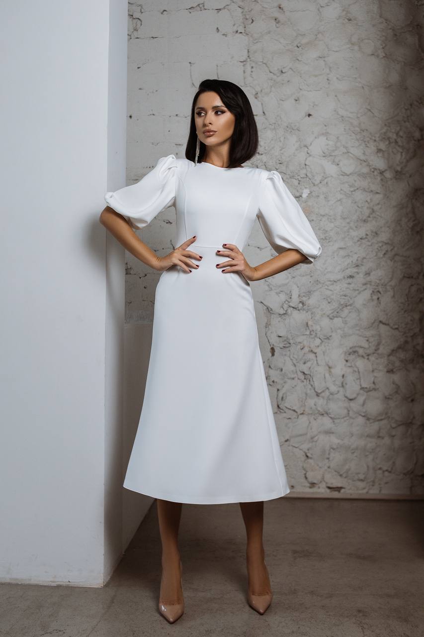 Woman wearing a white dress with puffed sleeves against a textured wall.