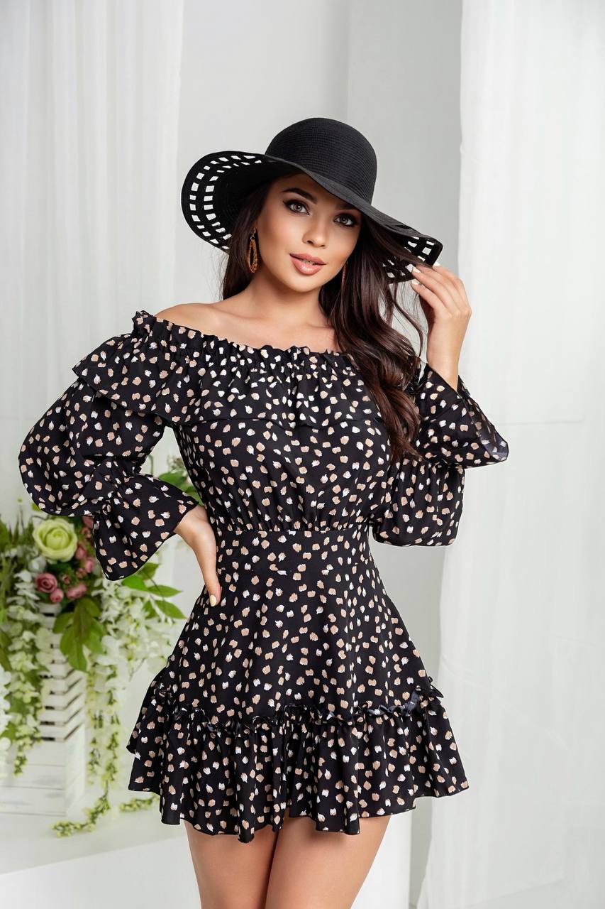 Woman wearing a black polka dot dress and black hat against a white curtain background