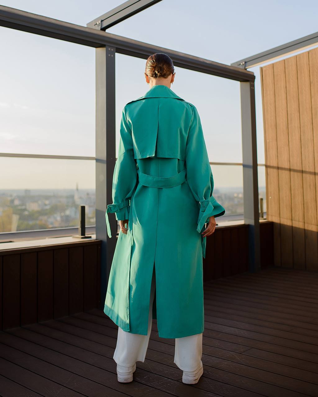 Person wearing a teal trench coat standing on a rooftop with a cityscape view.