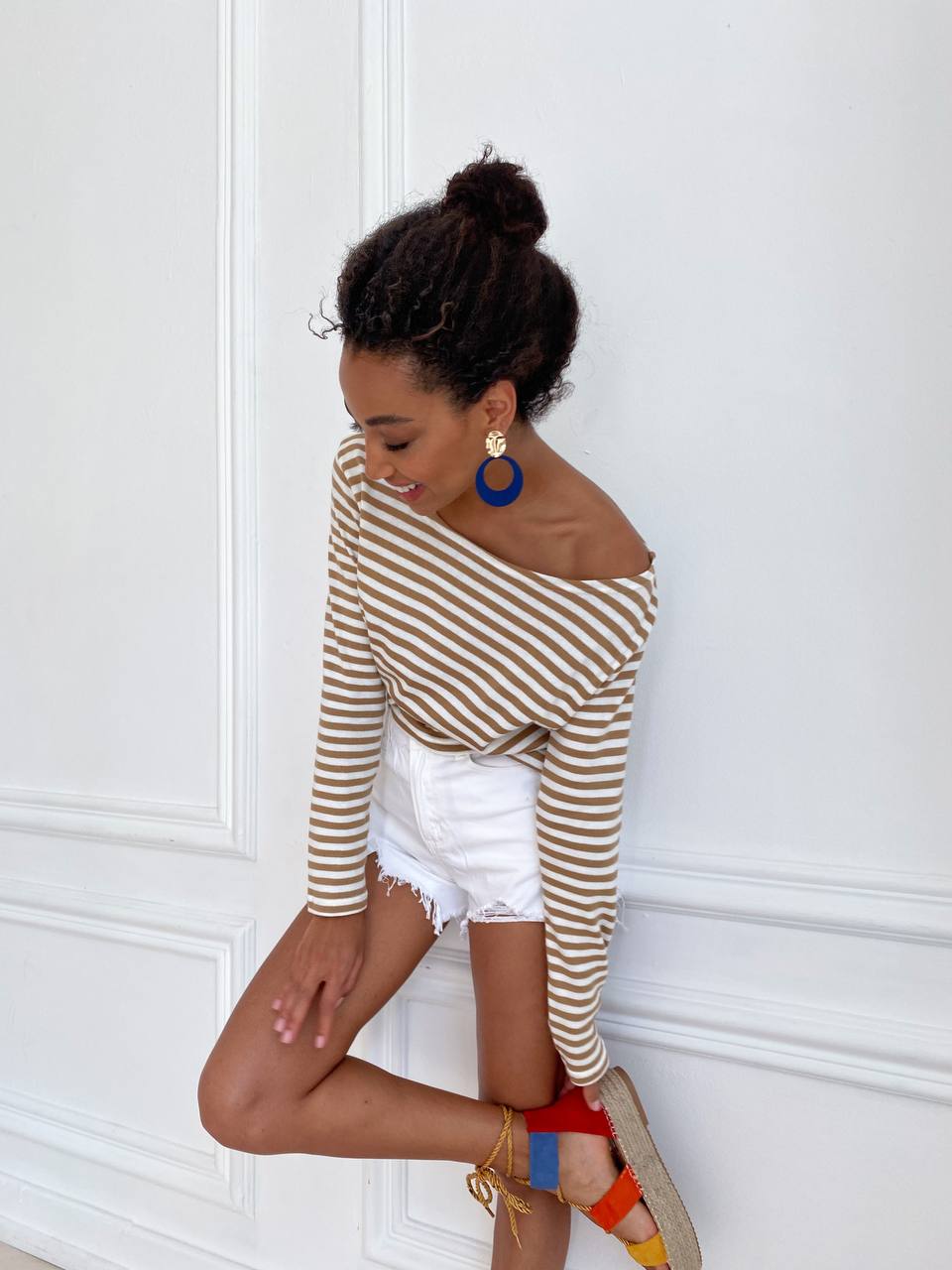 Woman wearing a brown and beige striped top with white shorts against a white wall.