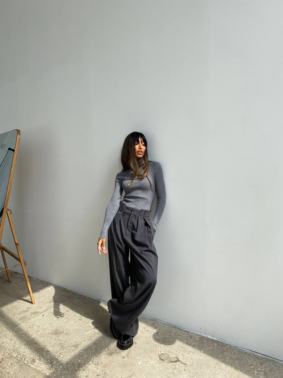 Person wearing a gray sweater and black pants standing against a white wall.