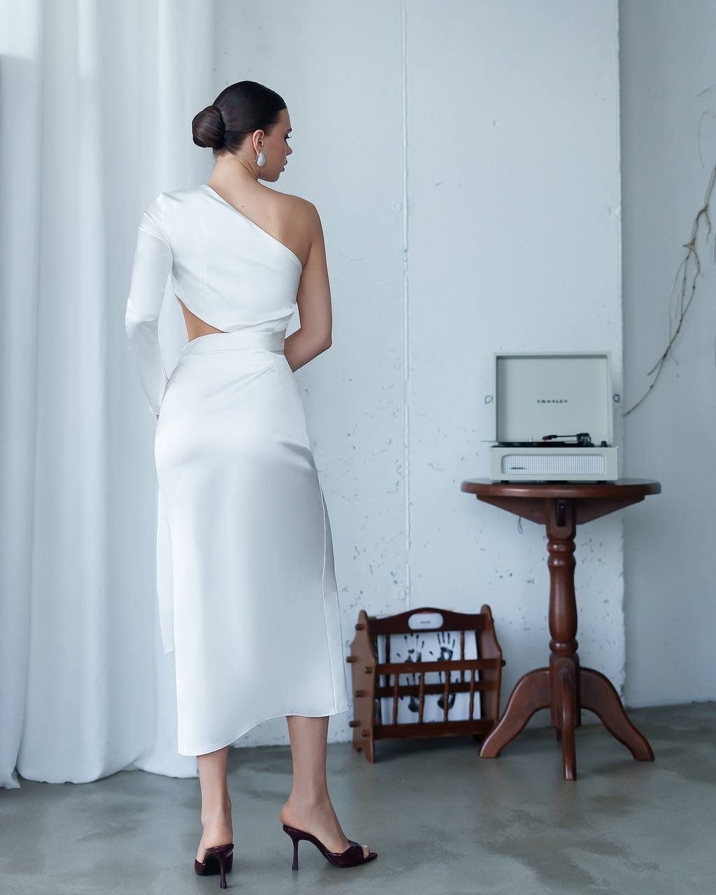 Woman in a white one-shoulder dress standing in a minimalistic room with a vinyl record player.