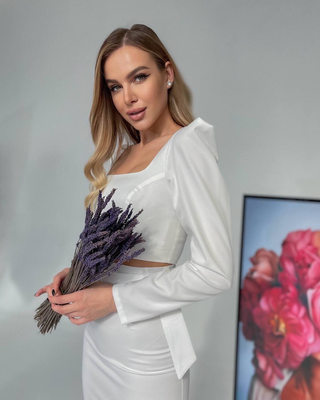 Woman in a white outfit holding lavender flowers against a neutral background