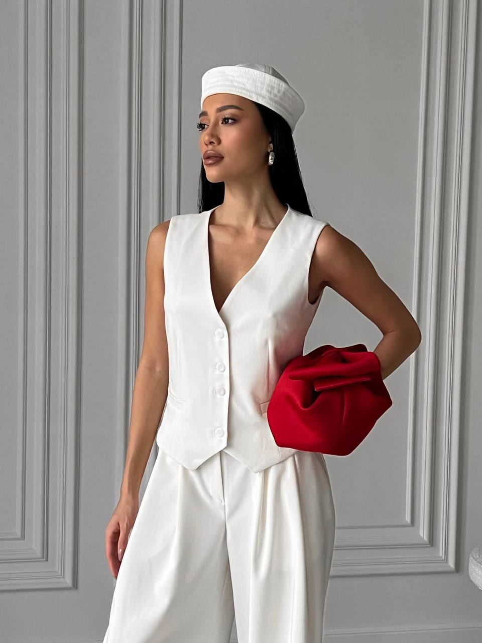 Woman in a white outfit holding a red clutch against a gray wall