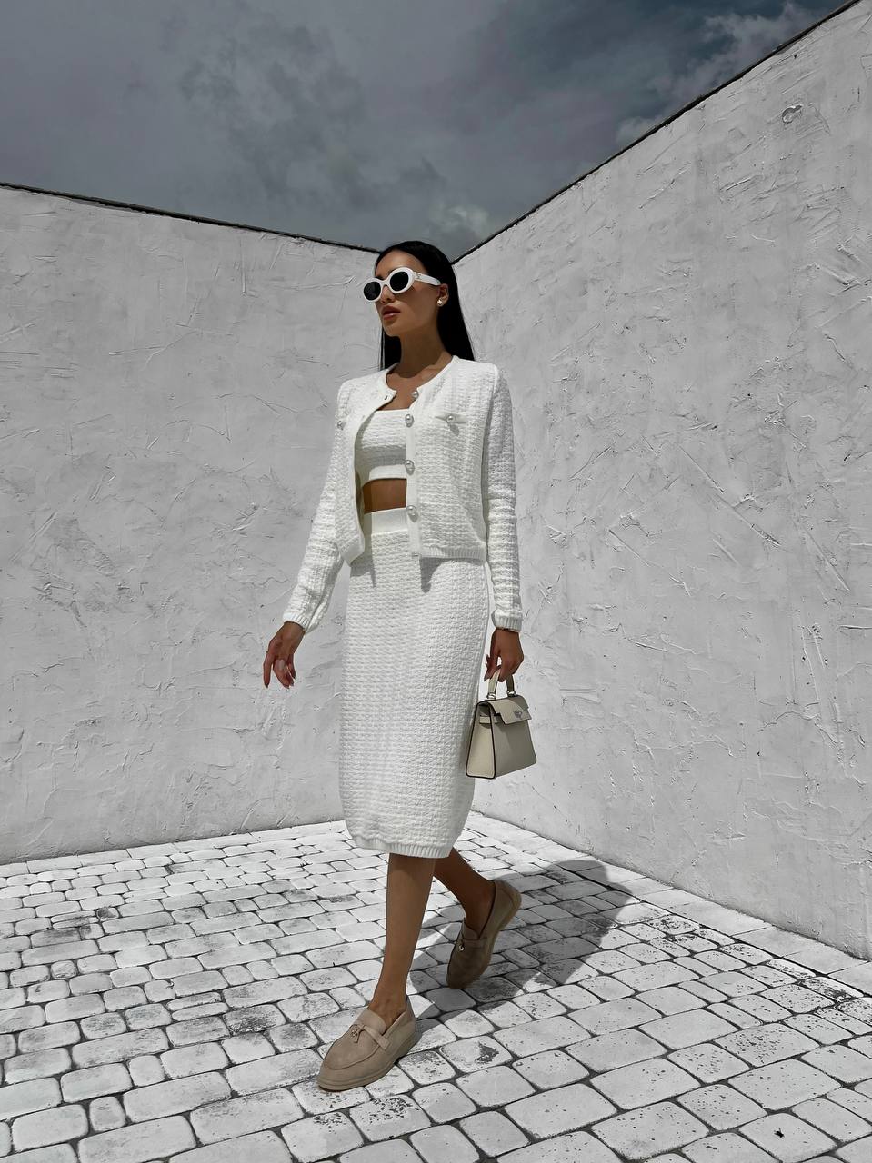 Woman in a white outfit walking in an open area with white walls and gray sky.
