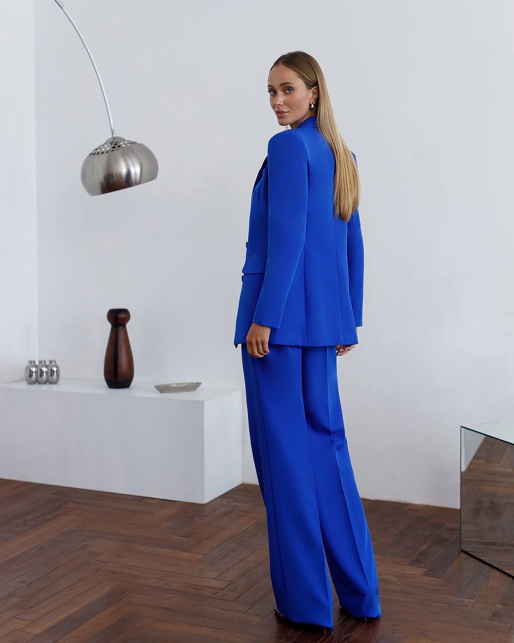 Woman in a blue suit standing in a modern interior setting with a white wall and wooden floor.