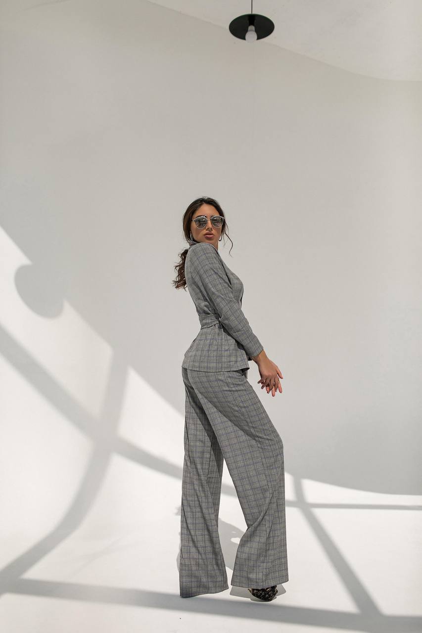 Woman wearing a gray checkered suit standing against a white wall with shadows.