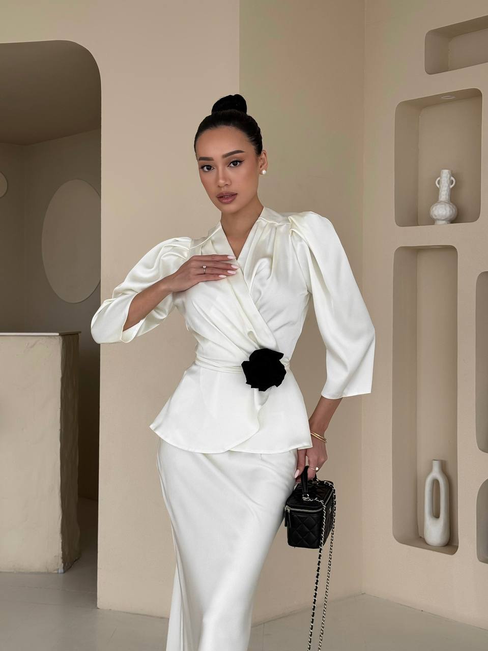 Woman in a white outfit with a black flower detail, holding a black handbag, against a neutral wall.