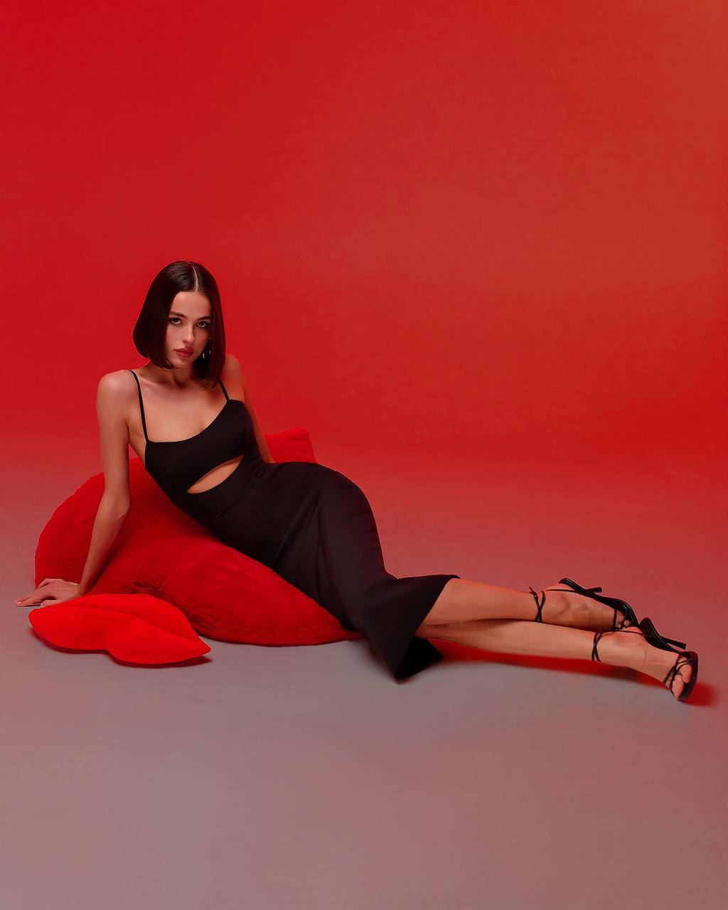 Woman in a black dress sitting on a red heart-shaped cushion against a red background
