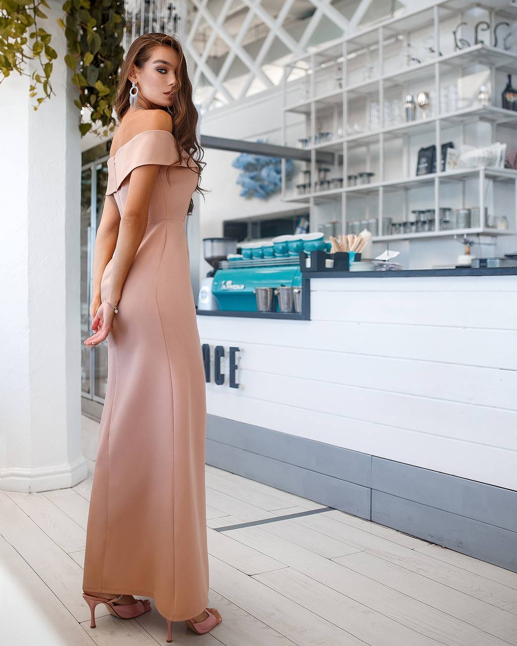 Woman in a long beige dress standing in a modern indoor setting with white walls and wooden floor.