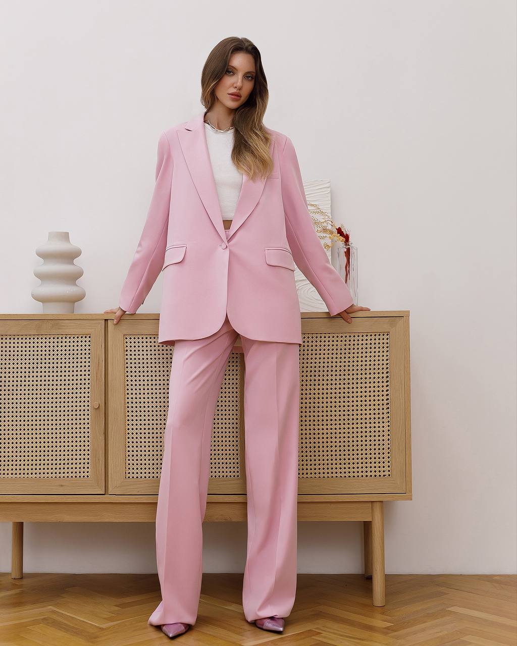 Woman in a pink suit standing against a light-colored wall with a wooden cabinet.