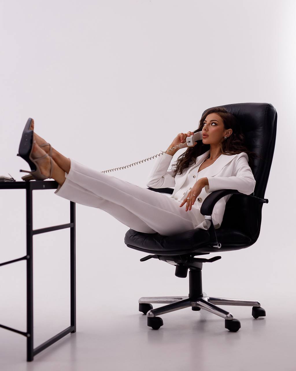 Woman sitting in a black office chair with feet on a desk, talking on a phone.