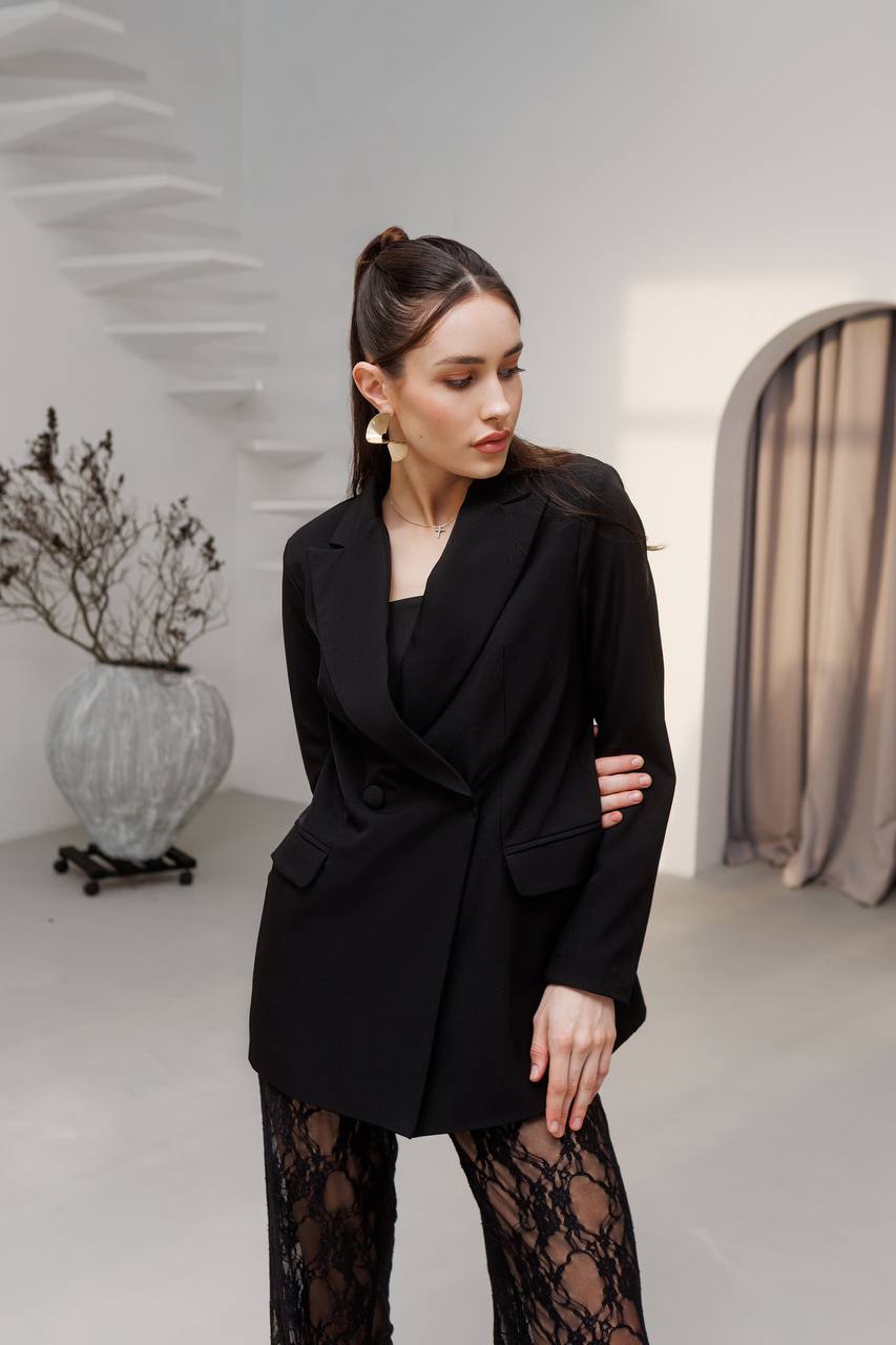 Woman in a black blazer and black lace pants standing in a minimalistic room.