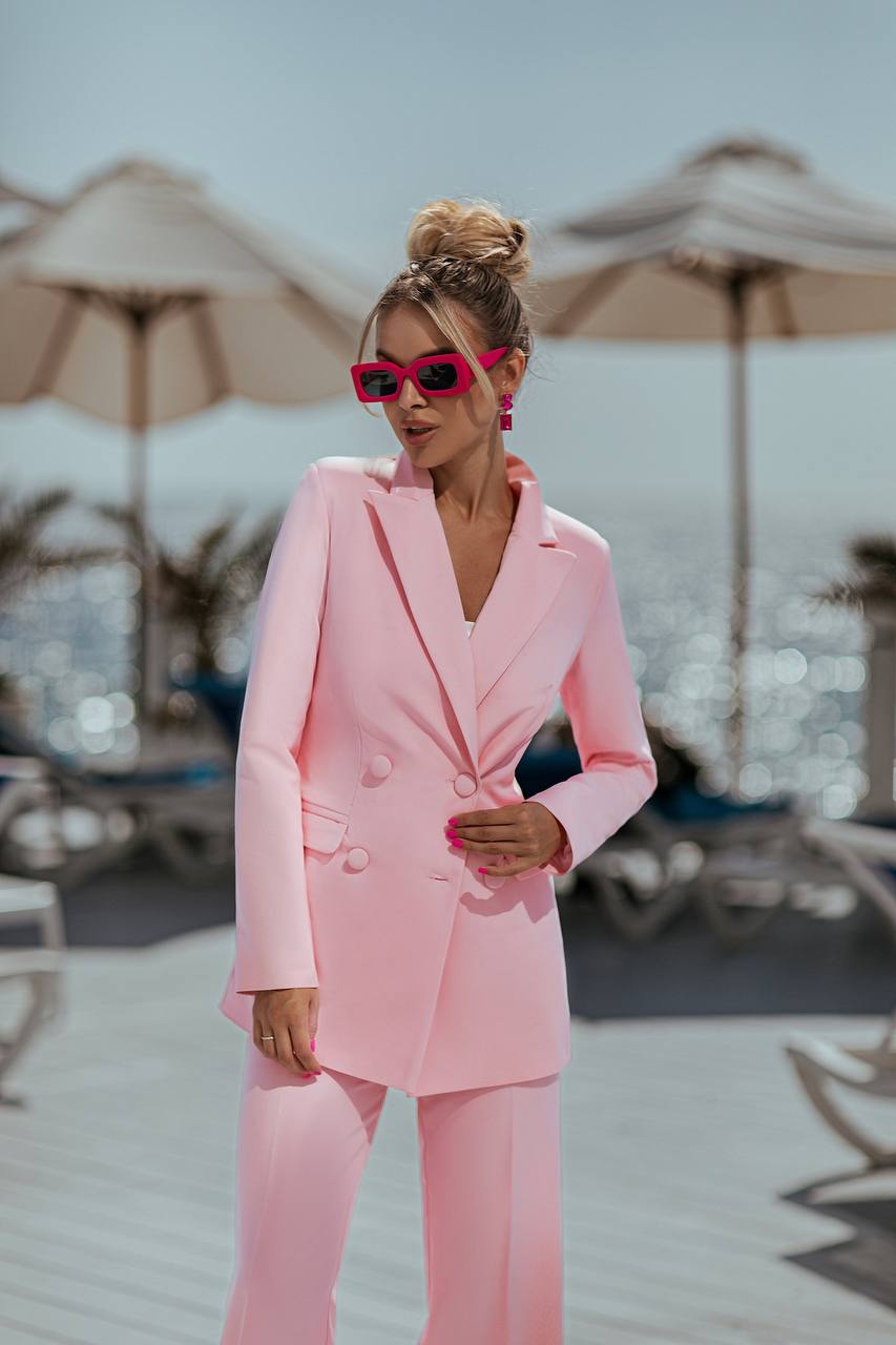 Woman in a pink suit standing on a beach with umbrellas and chairs in the background.