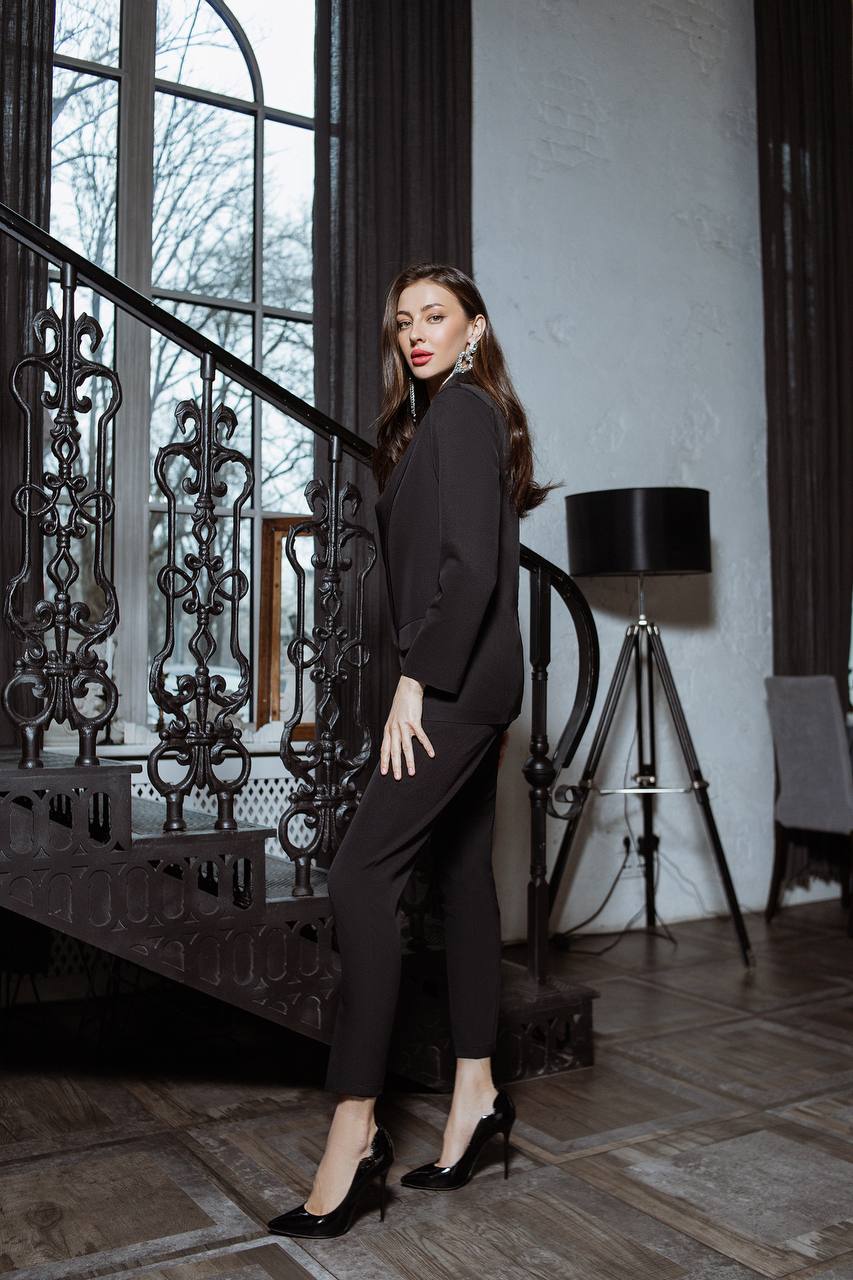 Woman in a black suit standing in an elegant interior setting with a staircase and lamp.
