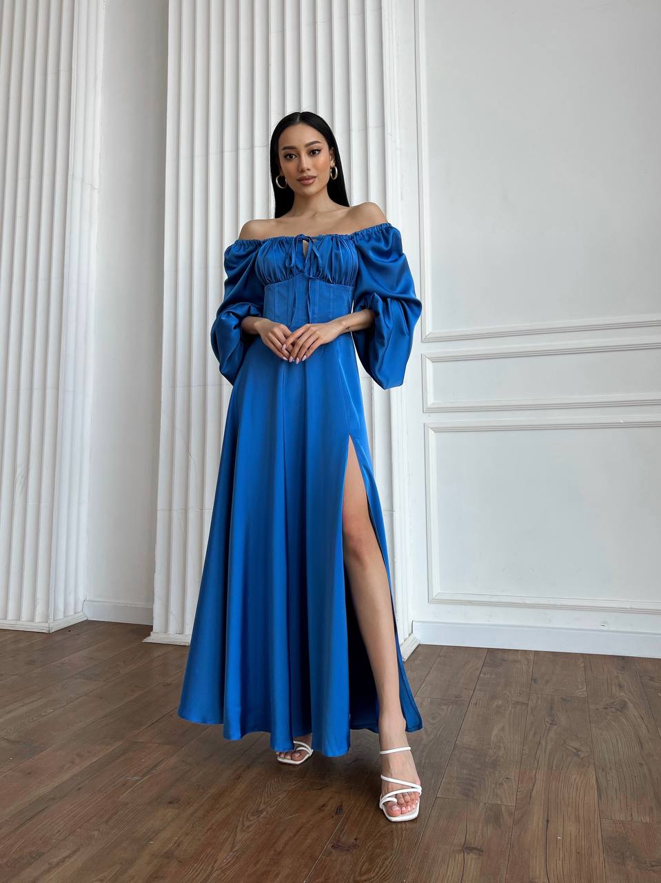 Woman wearing a blue off-shoulder gown with a high slit in a room with white walls and wooden floor.