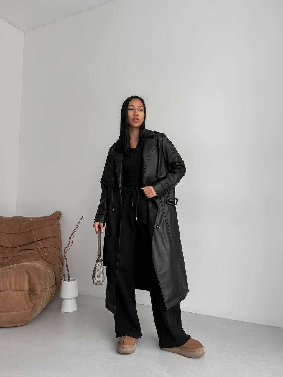 Person wearing a black trench coat in a minimalistic room with a brown sofa and white walls.