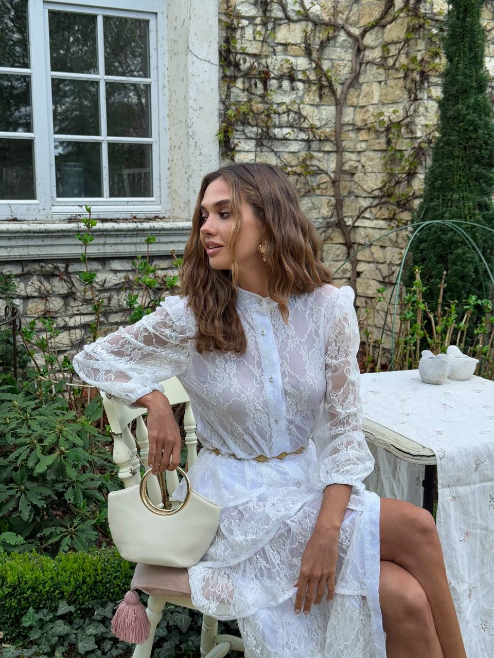 Woman in a white lace dress sitting outdoors with stone building and greenery in the background