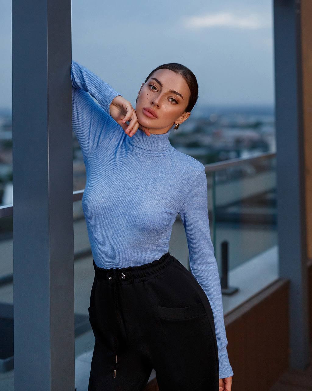 Woman in a blue sweater and black pants standing on a balcony with a cityscape view.