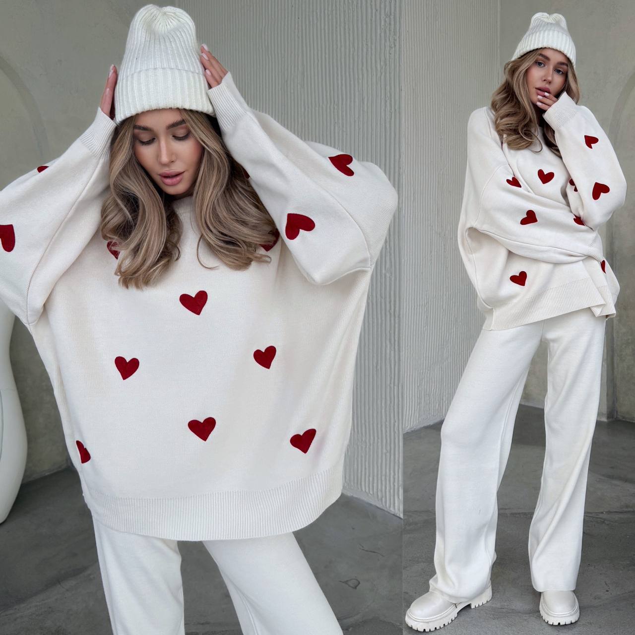 Woman wearing a white sweater with red heart patterns and white pants, standing against a neutral background.