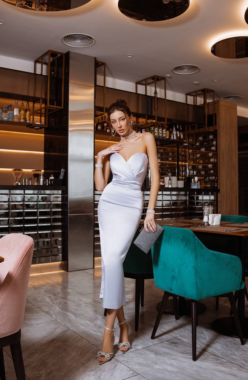 Woman in a white dress standing in a stylish interior setting with green chairs and a bar in the background.