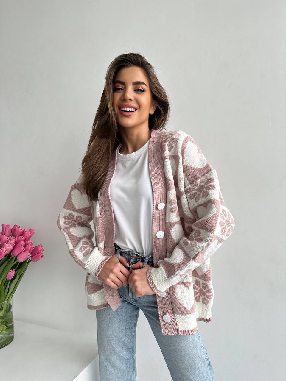 Woman wearing a patterned pink cardigan over a white shirt and blue jeans, standing against a plain background with a vase of pink tulips.