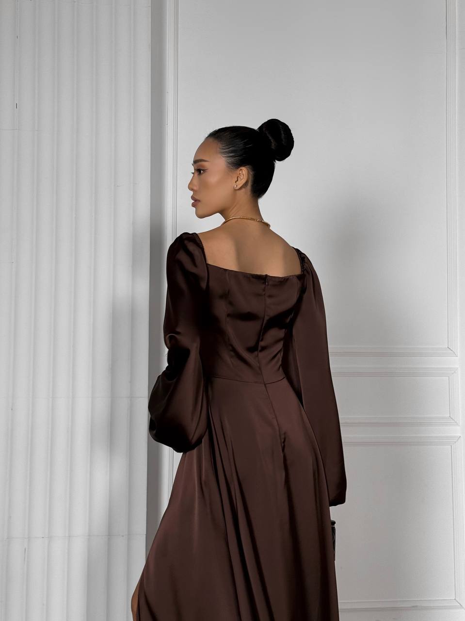 Woman in a brown dress standing against a white paneled wall.