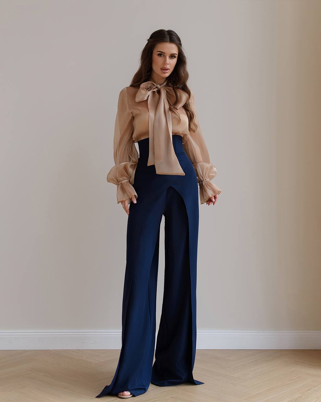 Woman wearing a beige blouse with a large bow and navy blue pants against a plain wall.