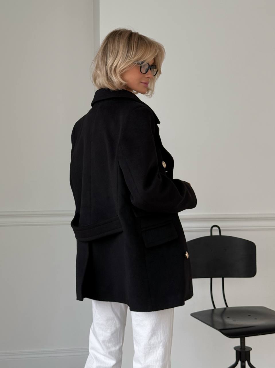 Person wearing a black coat and white pants in a minimal indoor setting