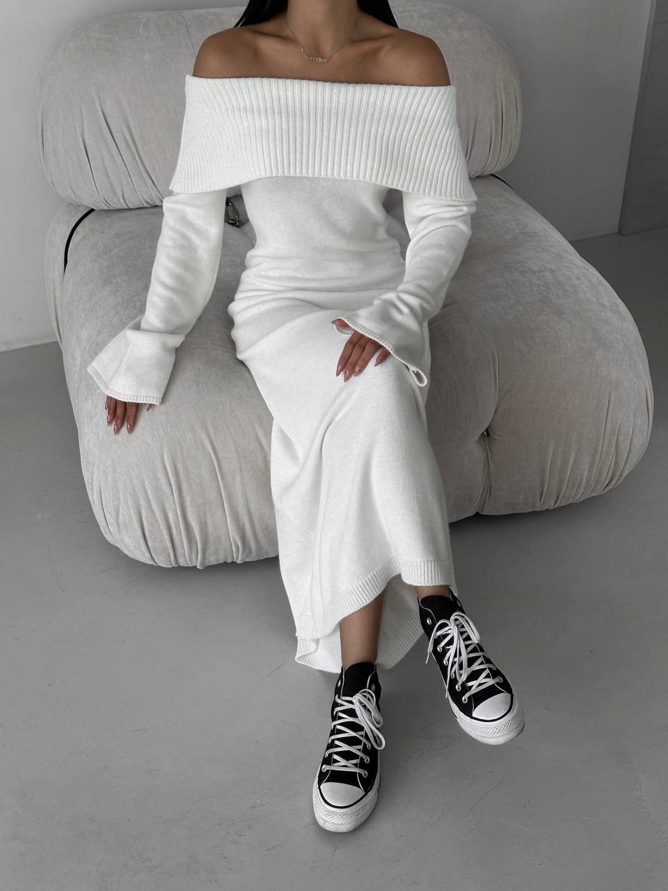 Person wearing a white off-shoulder top and long skirt with black high-top sneakers, sitting on a gray couch.