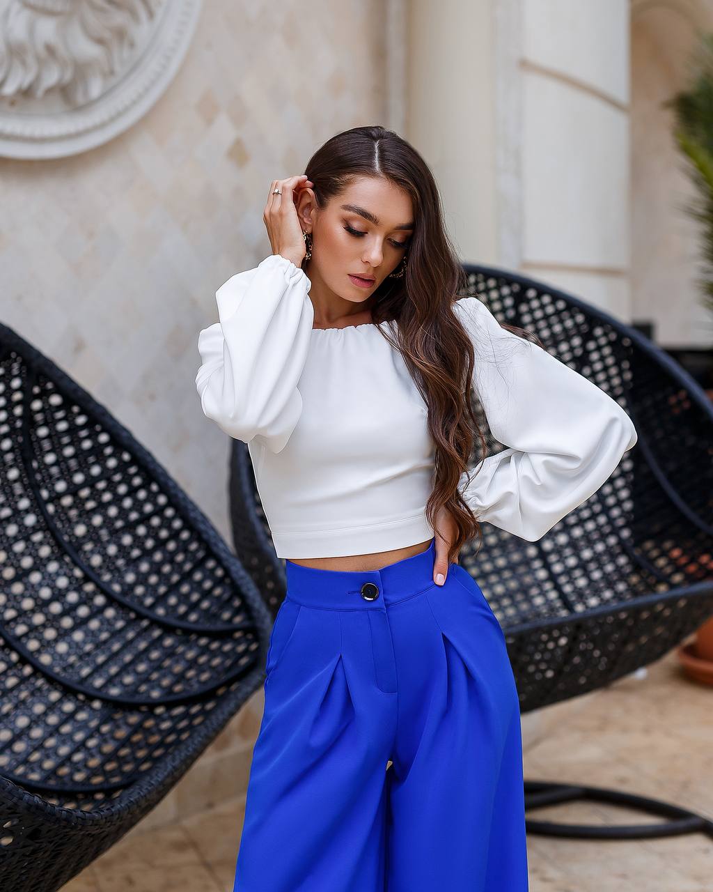 Woman wearing a white blouse and blue pants standing in front of wicker chairs.
