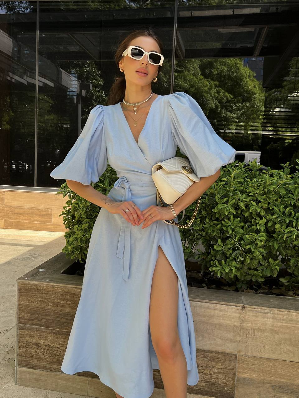 Woman in a light blue dress with a high slit, standing outdoors near greenery.