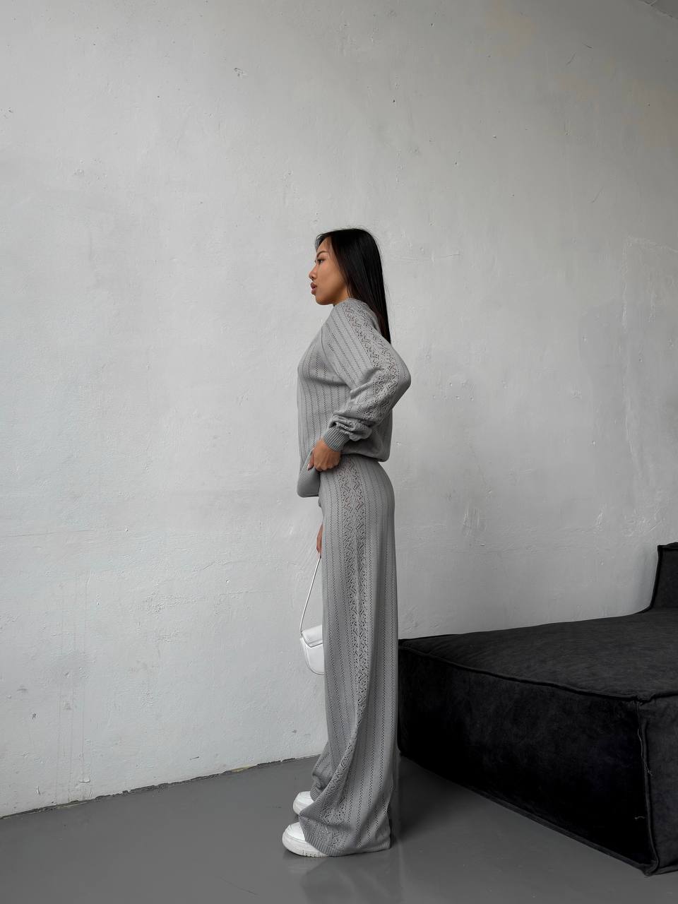 Woman in a gray knit outfit standing against a plain wall with a black couch.