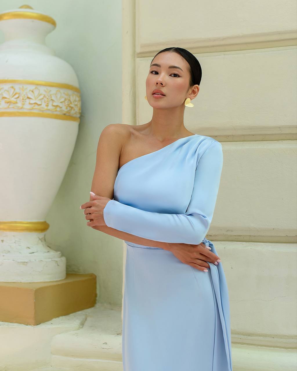 Woman in a light blue one-shoulder dress standing next to a decorative vase.