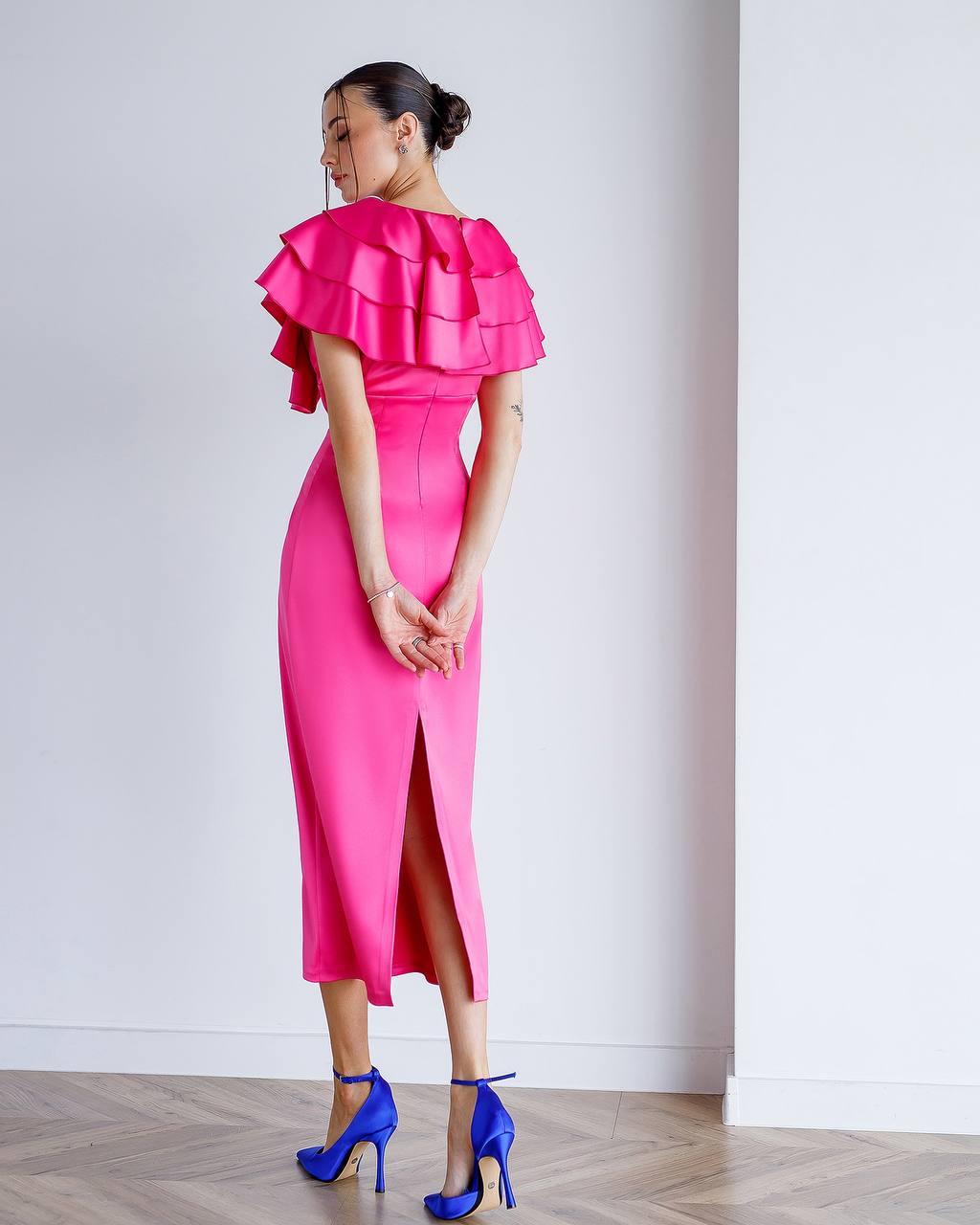 Woman wearing a bright pink dress with ruffled top and high slit, standing against a white wall.