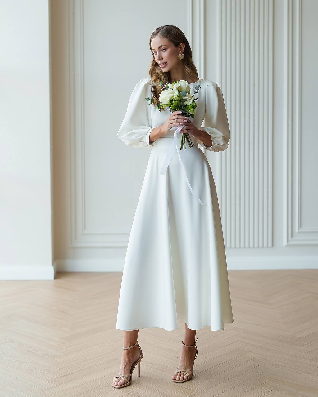 Woman in a white dress holding a bouquet in a room with wooden flooring and white walls.