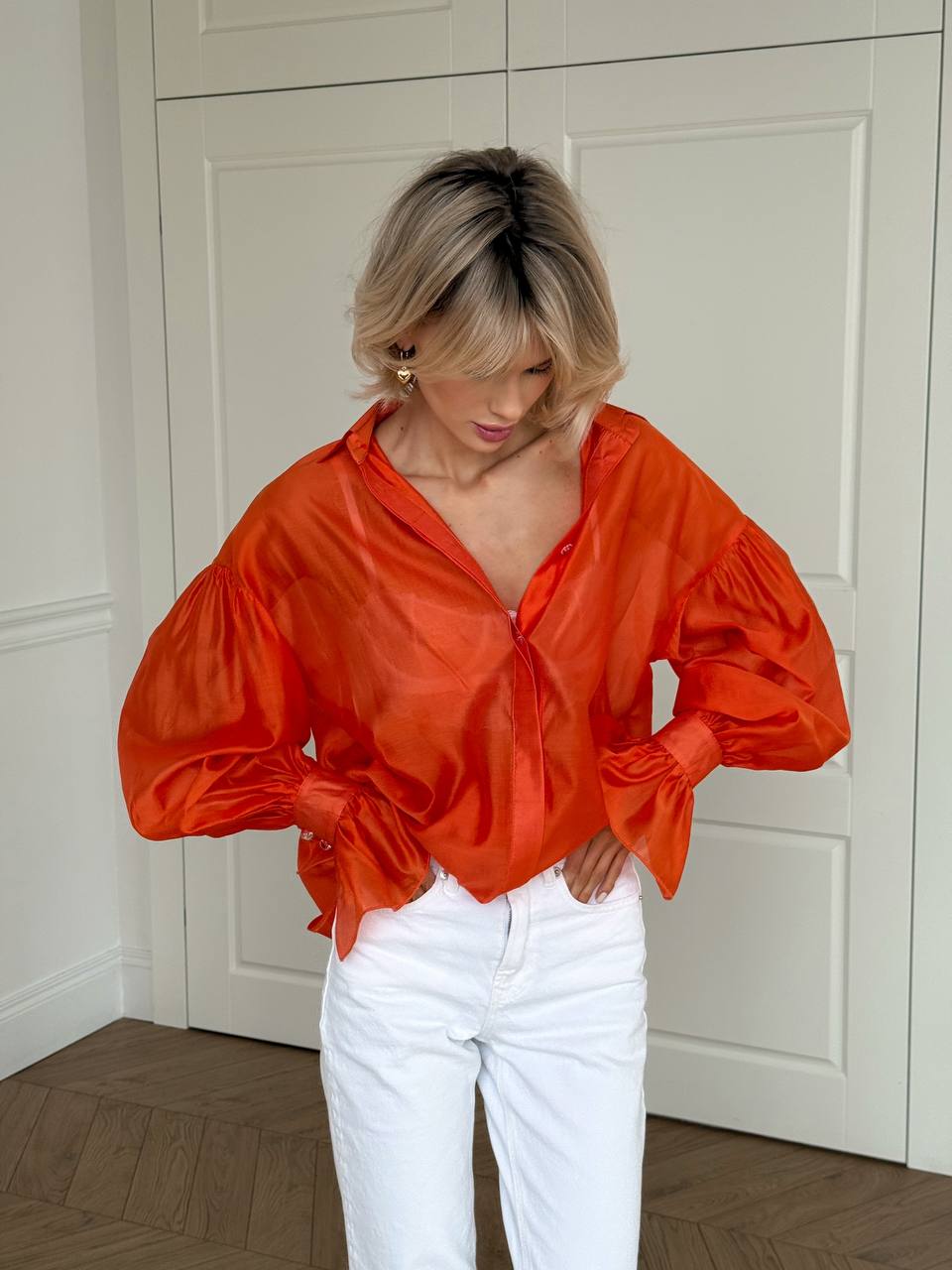 Person wearing an orange satin blouse and white pants in a room with white walls and wooden floor.