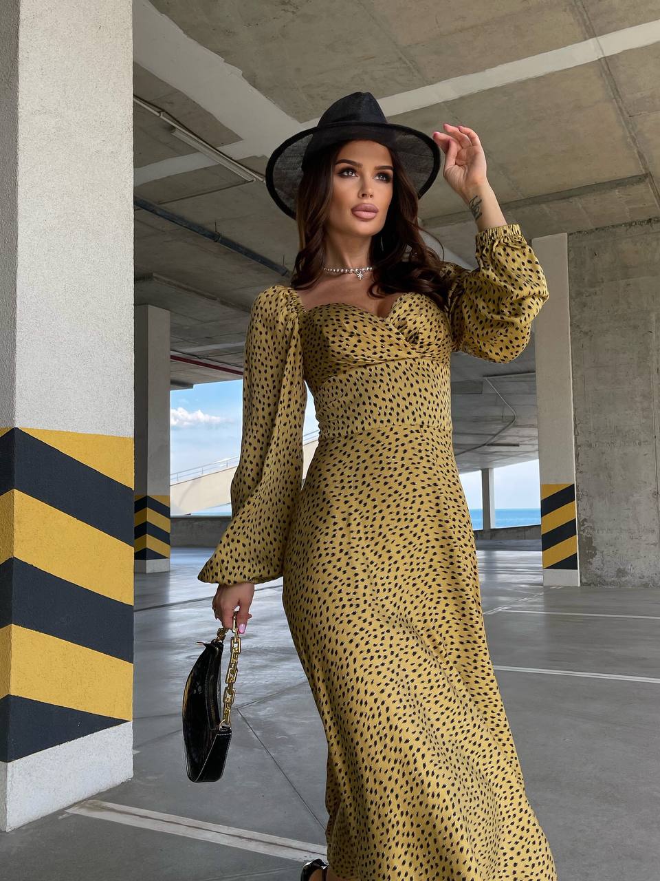 Woman in a yellow dress and black hat standing in an outdoor setting with concrete pillars and sky visible.