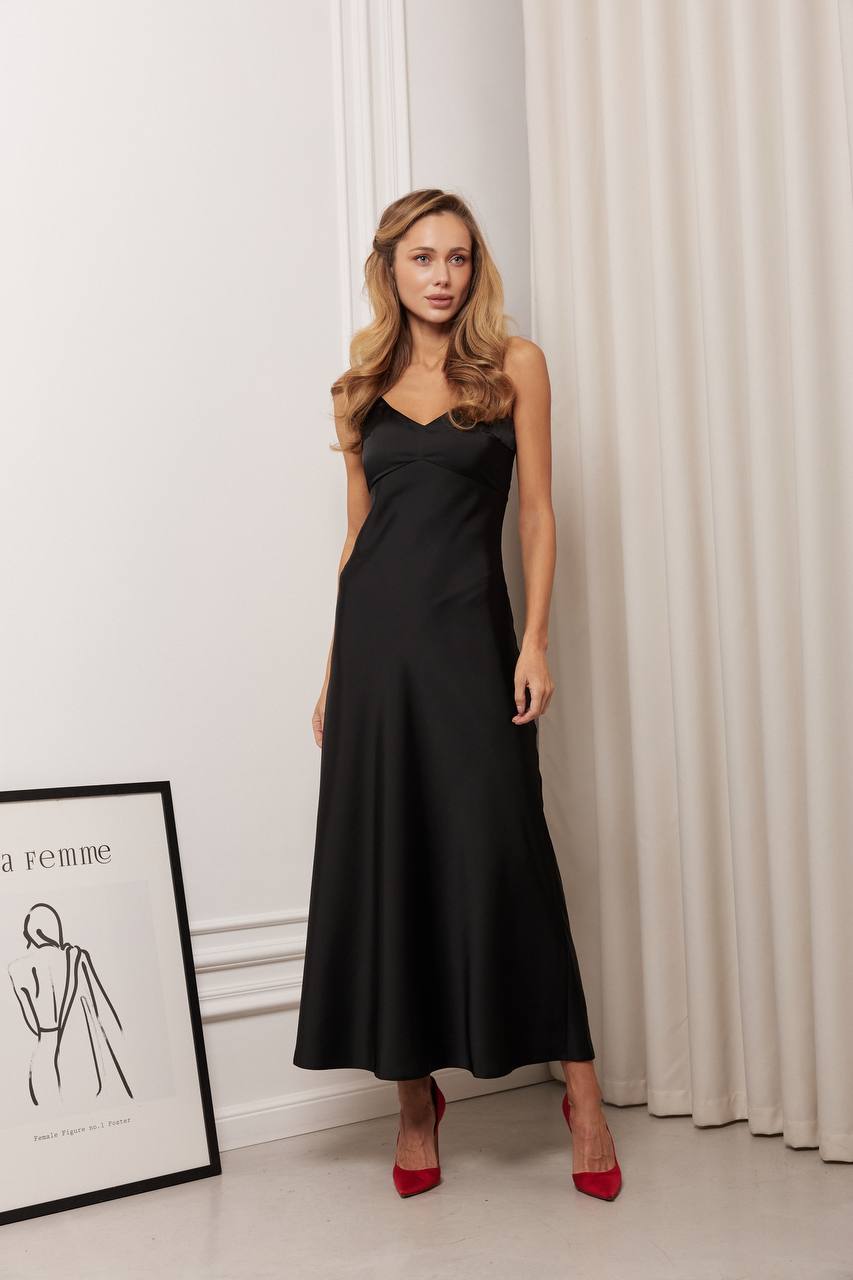 Woman wearing a black dress standing in a room with white curtains and a framed picture on the wall.