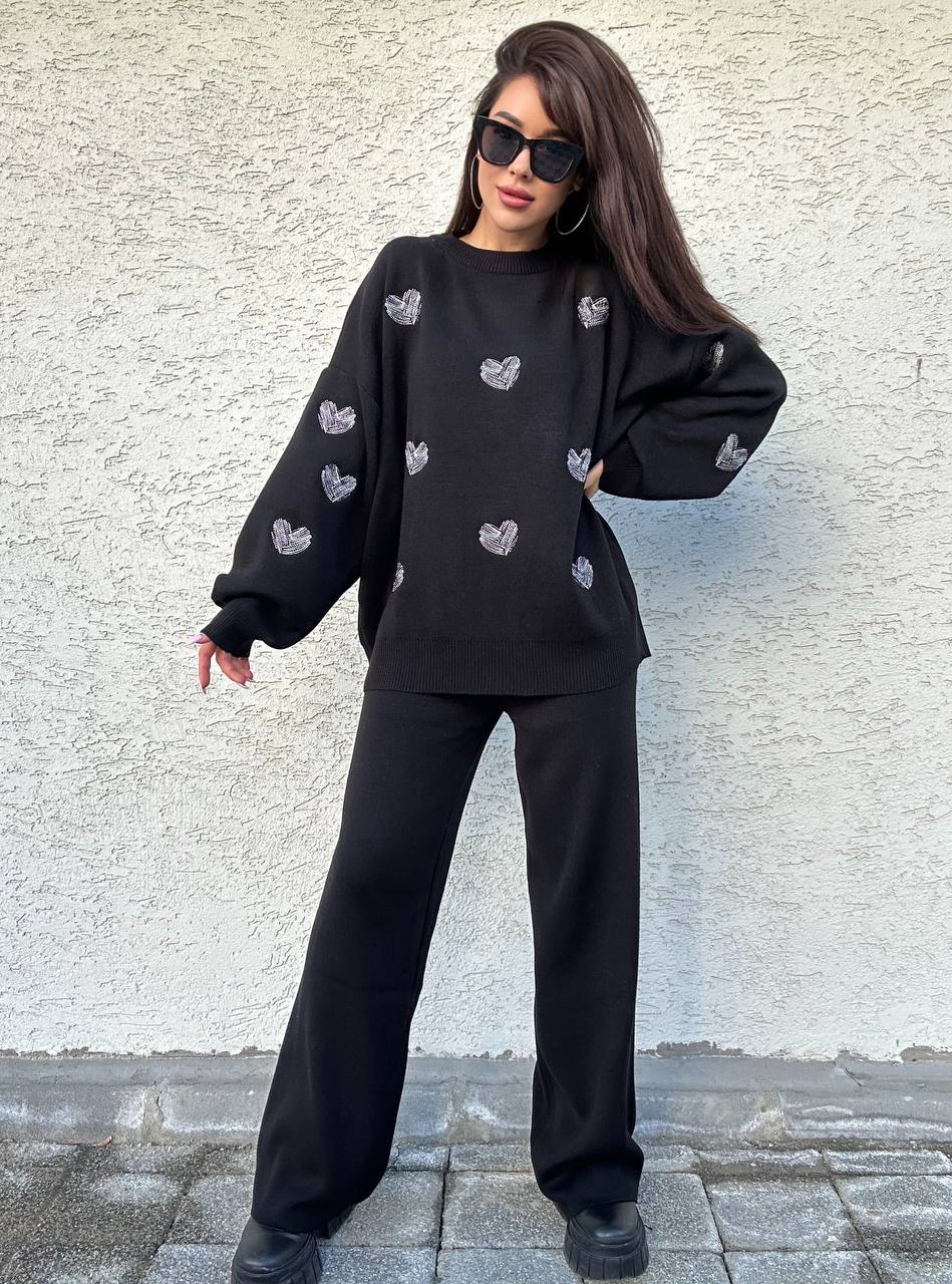 Person wearing a black outfit with heart patterns against a light gray wall.