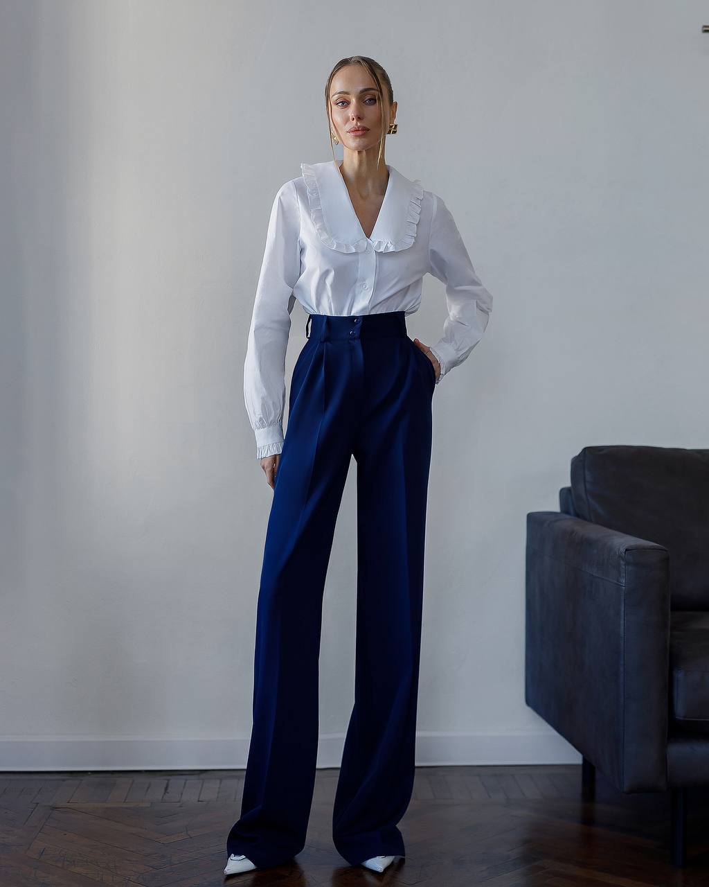Woman wearing a white blouse and blue pants standing against a plain wall.